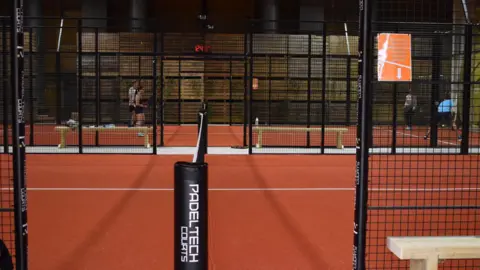 BBC/Hannah Sackville-Bryant We can see two padel courts - our viewpoint is in line with the net. They resemble tennis clay courts and are surrounded by metal meshed fencing.

The nearest court is empty, the far one has four players on it.