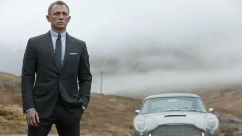 Danjaq LLC/United Artists/Columbia Pictures Daniel Craig as 007 in Skyfall. He is on the left wearing a grey suit, with one button done up, over a light coloured shirt and dark tie. He has short fair hair and is brooding. On the right is a silver coloured car. Behind is Scottish scenery of heathery moorland and a grey misty sky. 