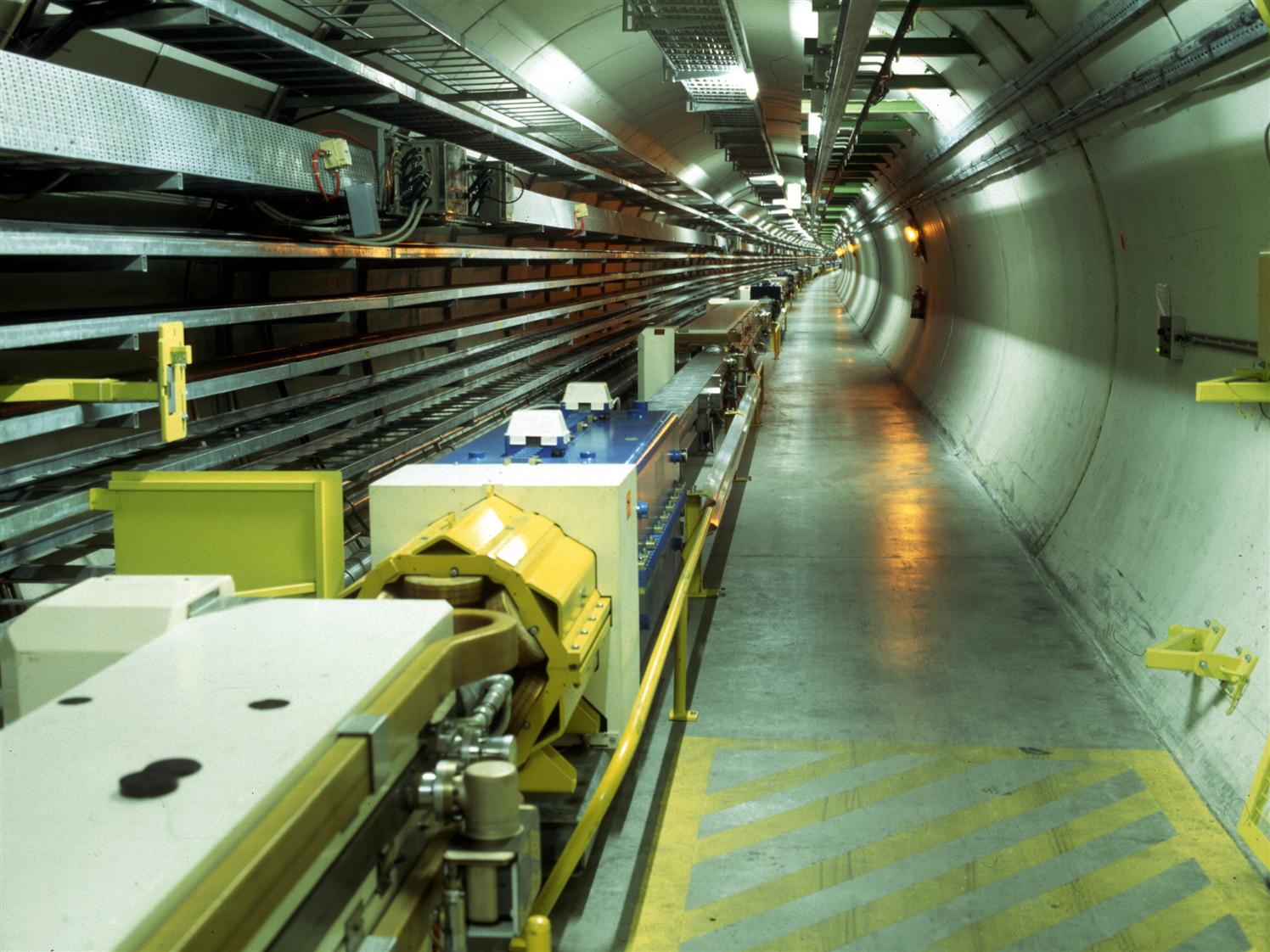 A long underground tunnel housing scientific equipment and cables, likely part of a particle accelerator facility or Higgs factory, with concrete floors and bright overhead lighting dedicated to advanced particle physics research.