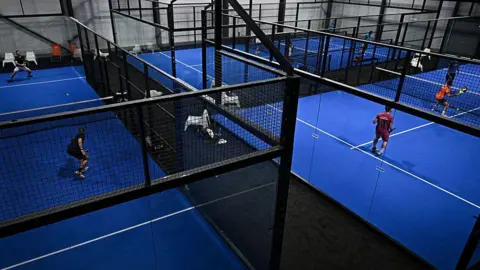 Getty Images We can see several padel courts at an indoors sports facilty. The courts are blue with white lines marked on them - and are surrounded by black, metal meshed fences. People are playing on them.