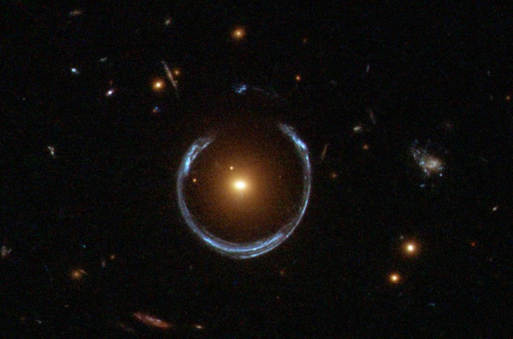 A bright galaxy at the center, possibly hiding the heaviest black hole, bends light from another galaxy behind it to form an Einstein ring—a striking blue circle surrounded by stars and galaxies.