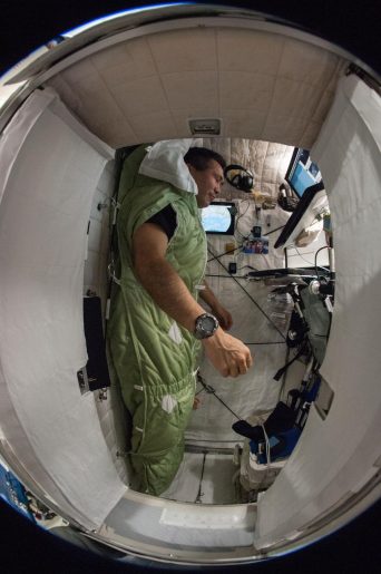 An astronaut inside a spacecraft, standing upright in a green sleeping bag-like garment. The compact interior features equipment and controls on the walls and ceiling, with a small window in front letting in light. The image has a fisheye lens effect, creating a slightly distorted perspective.