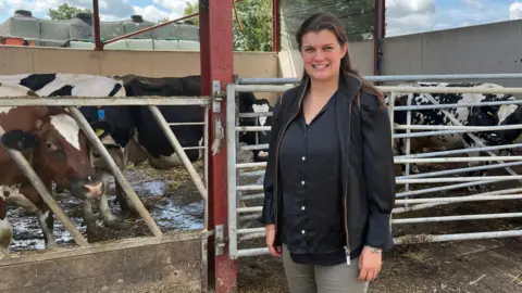 A young woman with long brown hair smiles as she stands in front of a shed full of cows. She is wearing a black shirt, a black waistcoat, and grey trousers.