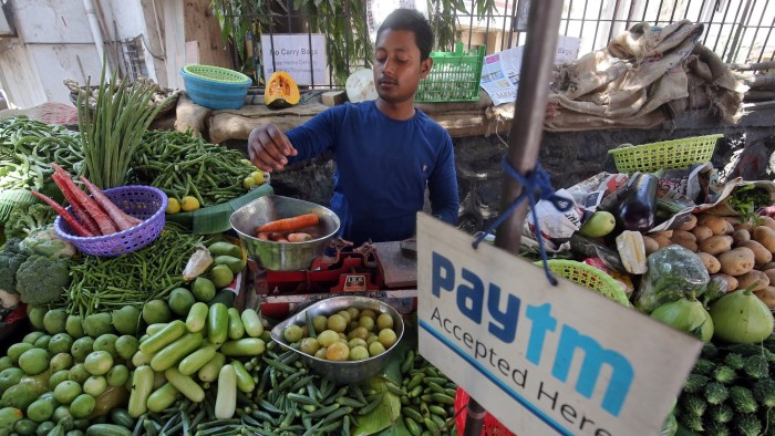 A vendor in a blue shirt weighs vegetables at a roadside market in Mumbai. A Paytm advertisement, indicating acceptance of digital payments, is prominently displayed among the baskets of fresh produce, which include carrots, potatoes, and various greens