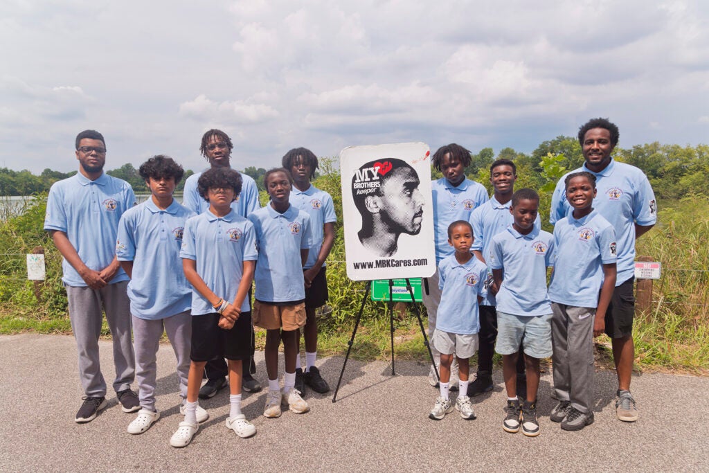 A number of Black male youths gather with instructors for a photo, standing alongside a poster