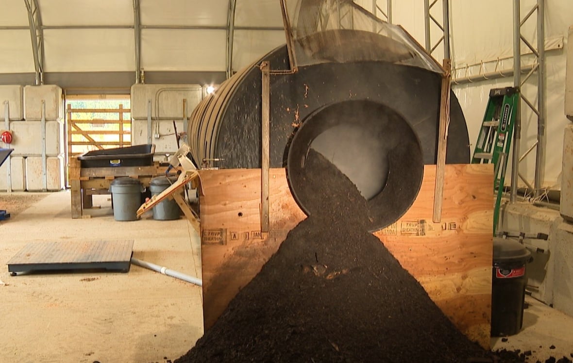 One end of a large black tube is seen with hot steam coming out and dark compost. The tube is inside a open-air barn.
