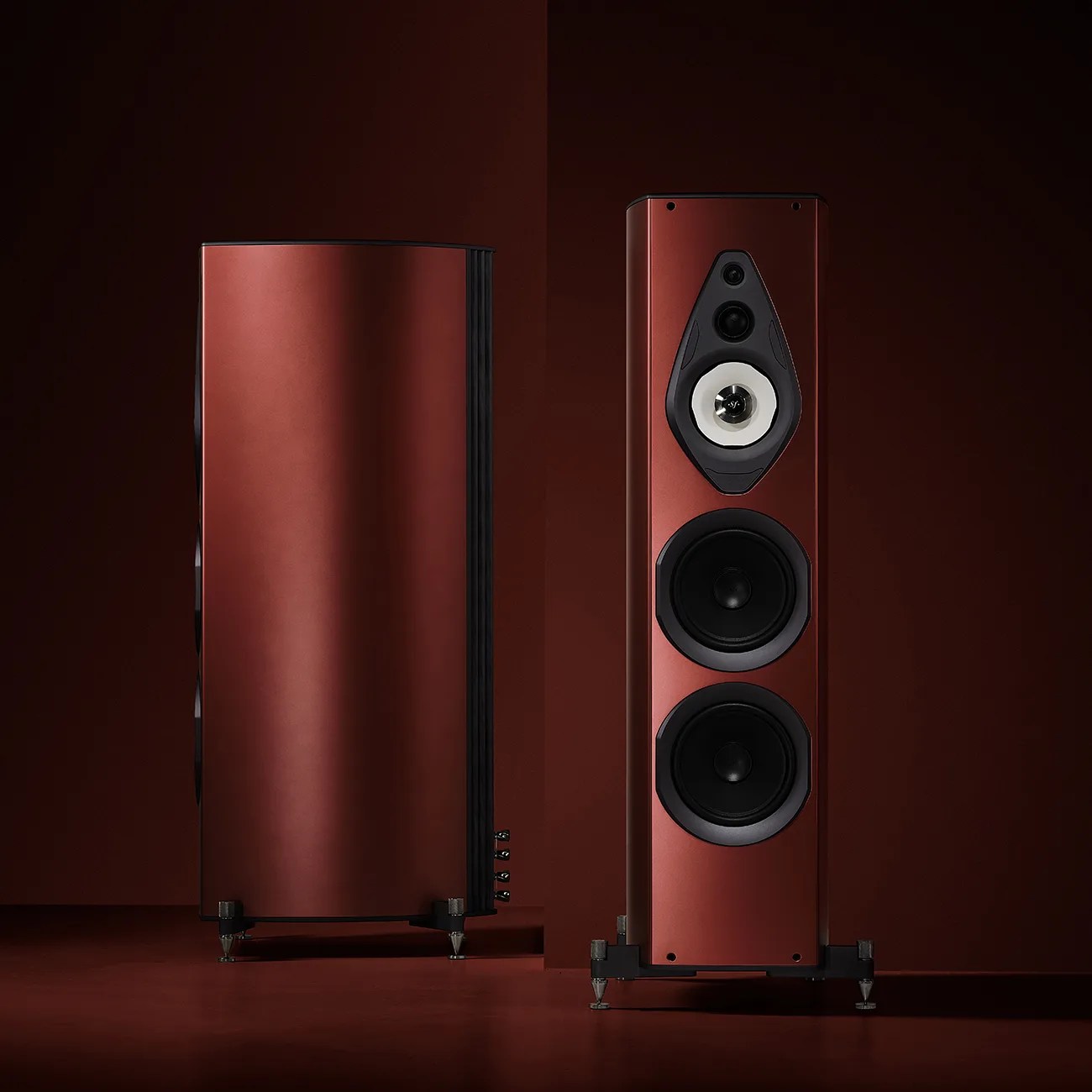Two tall, floor-standing speakers with a sleek, curved design and a matte reddish-brown finish. The front speaker shows three visible drivers: a small tweeter at the top, a midrange driver with a white cone below it, and two larger black woofers beneath. The speakers stand on metal spikes for stability, set against a matching reddish-brown background.