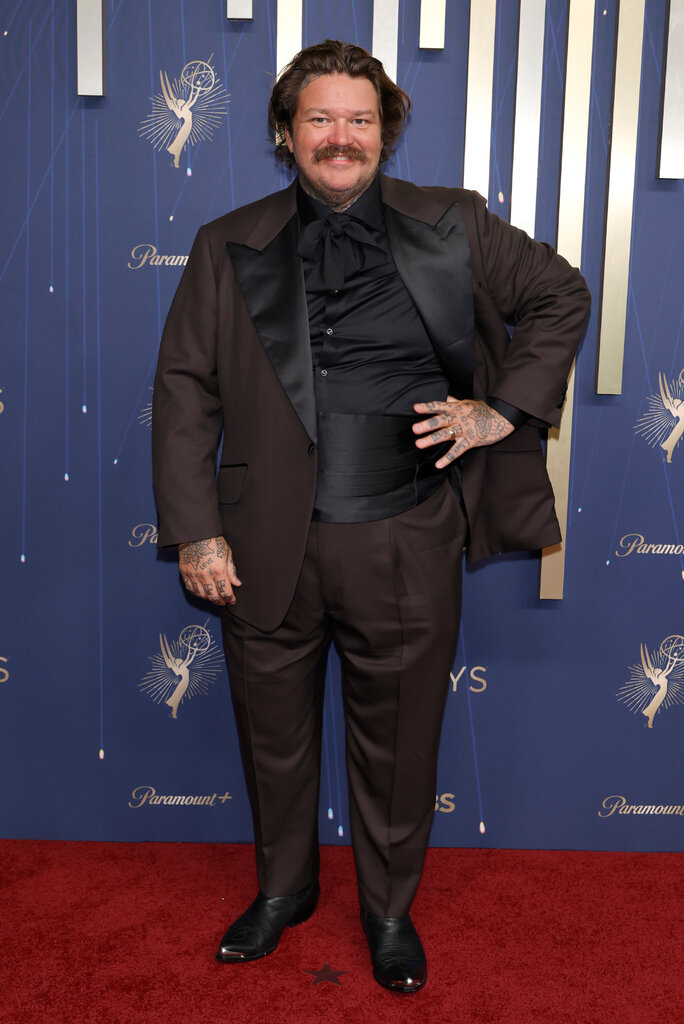 A man in a brown tuxedo with black details poses with his hand on his hip.