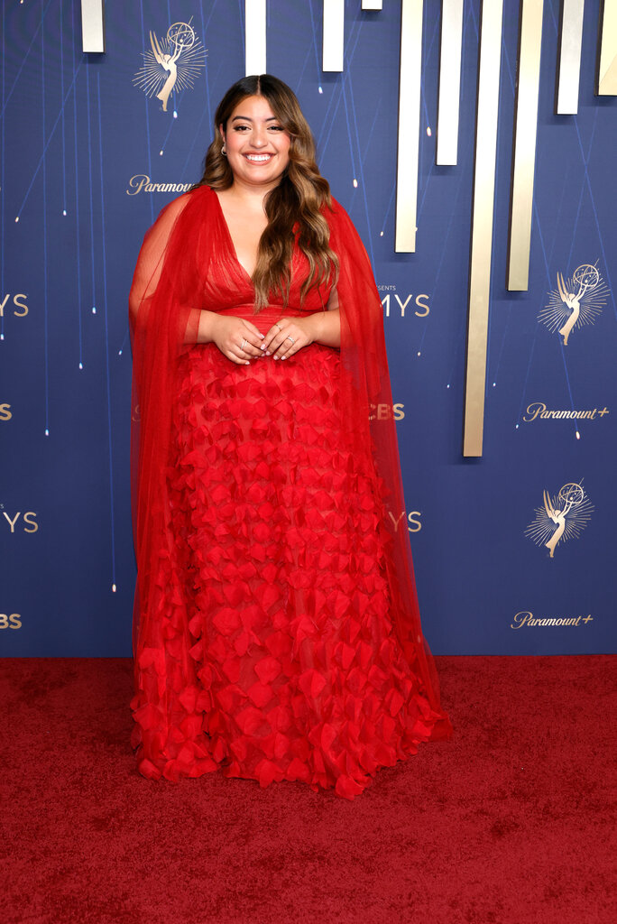 A woman in a red ruffled dress and matching sheer shawl poses on the red carpet.