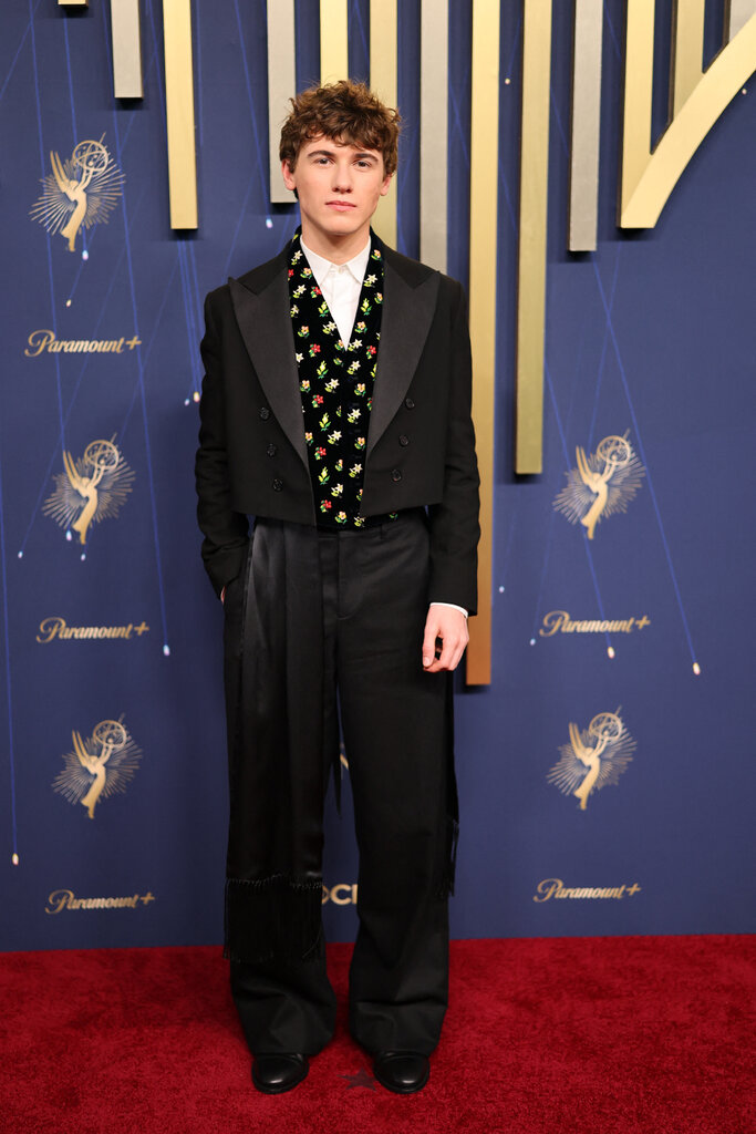 A man in a tuxedo with a floral vest poses with one hand in his pocket.