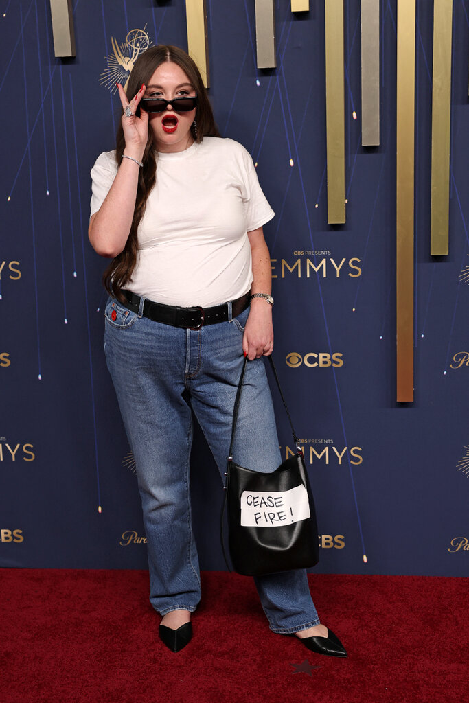 A woman poses with her mouth open and a hand lowering her sunglasses. She is wearing jeans and a white T-shirt and holding a bag that says "CEASE FIRE!" on it.