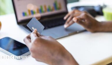 A person's holds a bank card in their left hand, while using a laptop trackpad with their right. On the computer screen are some bar charts.