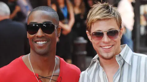 Getty Images Simon and Duncan stand smiling, each in a pair of sunglasses with a blurred crowd behind then. Simon wears a red T-shirt with a black lace, Duncan wears a white, grey and peach vertically striped shirt. Both have facial hair. 