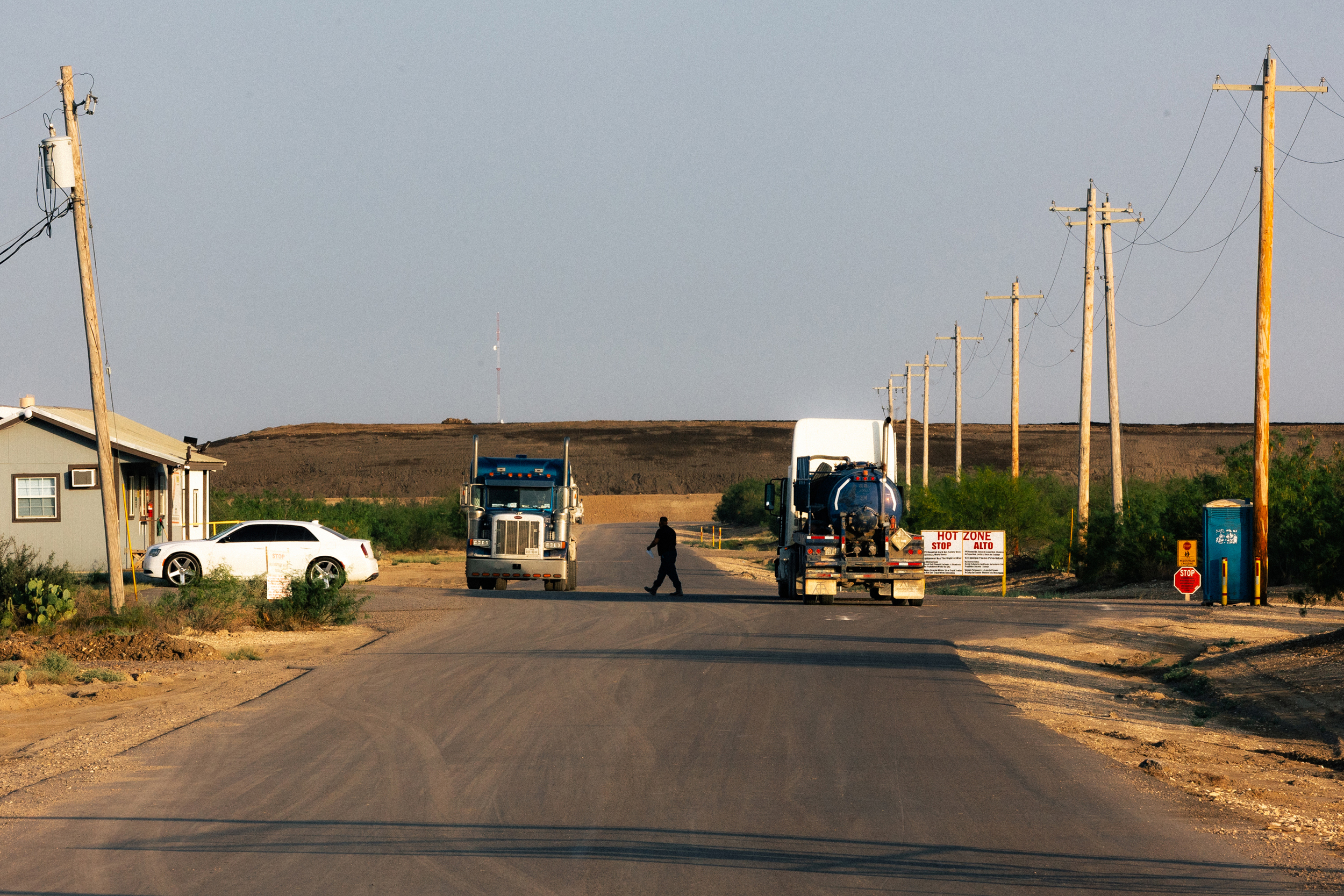 A man walks between two semitrucks on a road where only one small building is visible. A large, flat-topped hill made of darker soil stretches across most of the background and continues off the right side of the photo.