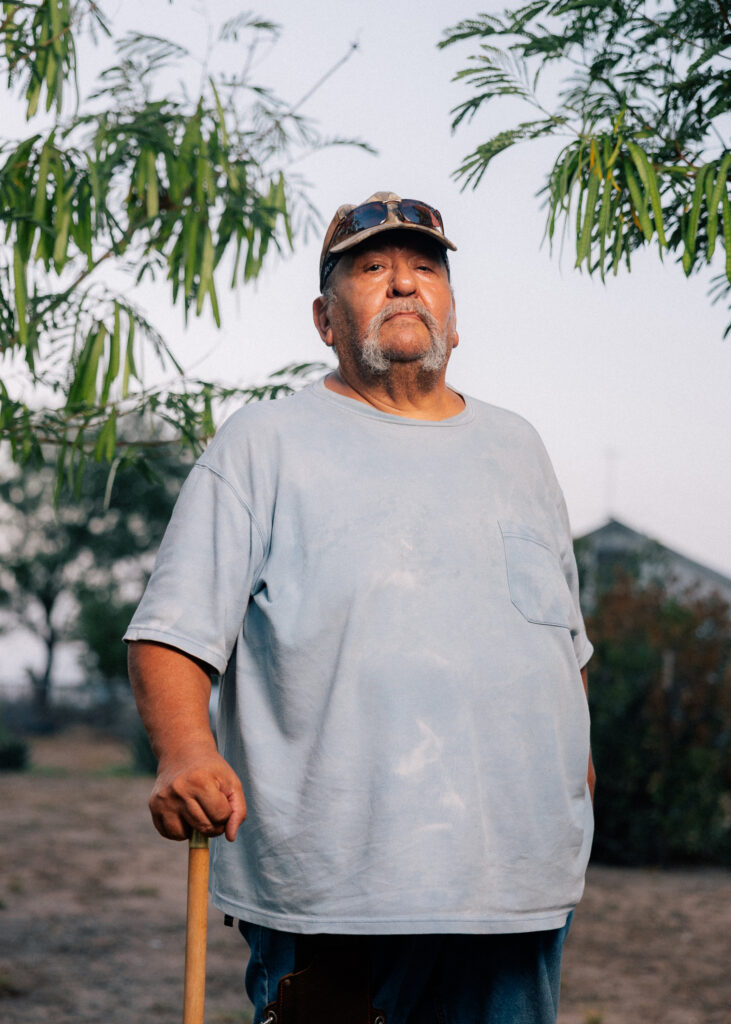 A man with a horseshoe mustache leans on a light wooden cane while standing outside. He wears a baggy blue T-shirt and a baseball cap with sunglasses resting on the brim.