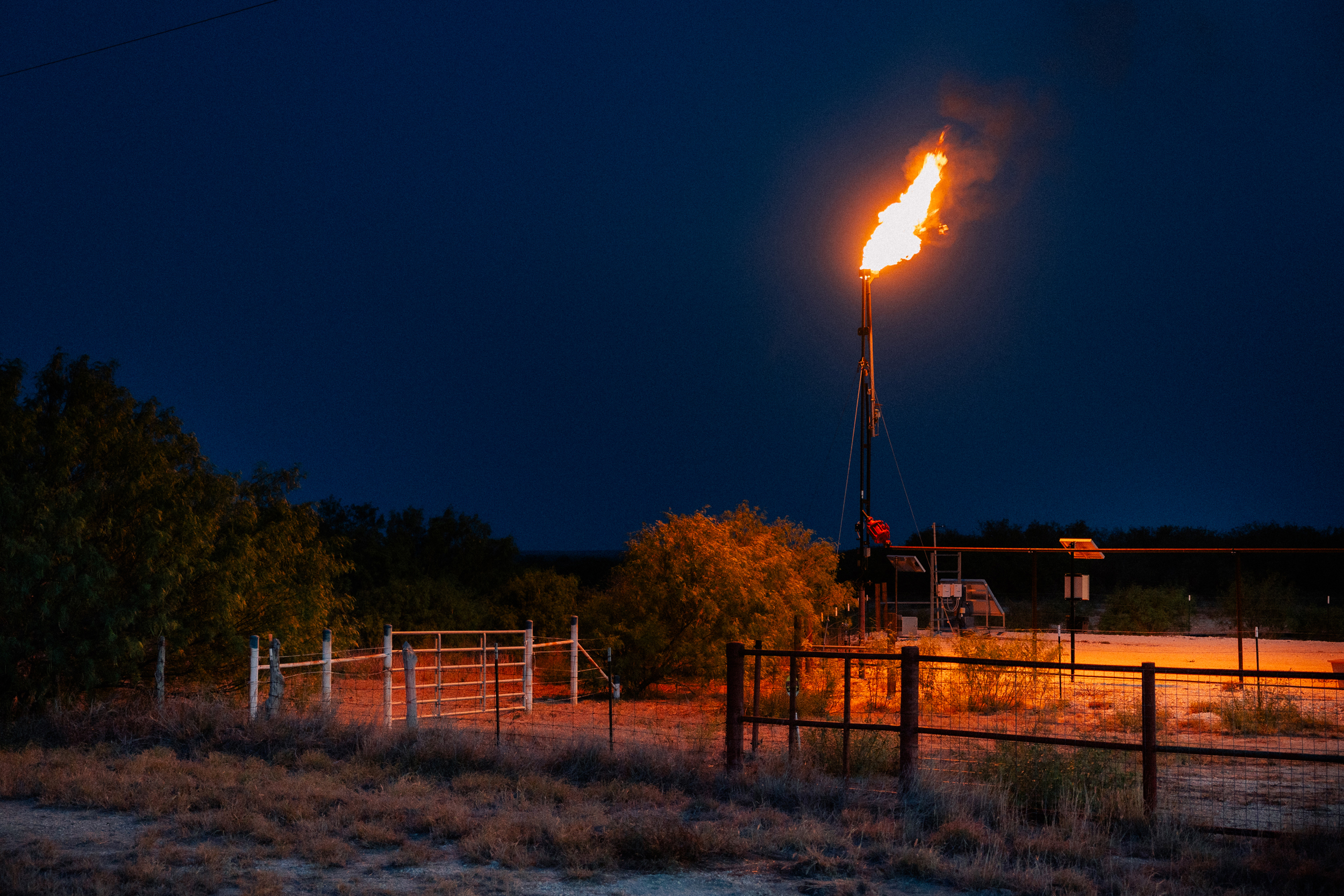A jet of flame rising from a tall pipe casts an orange glow over an area with fences and brush.