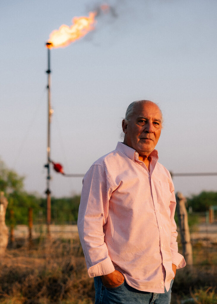 A man in a button-down shirt with thin pink and white vertical stripes stands with his hands in his jeans pockets. Behind him, flame jets from the top of a pipe that stands straight up in the air.