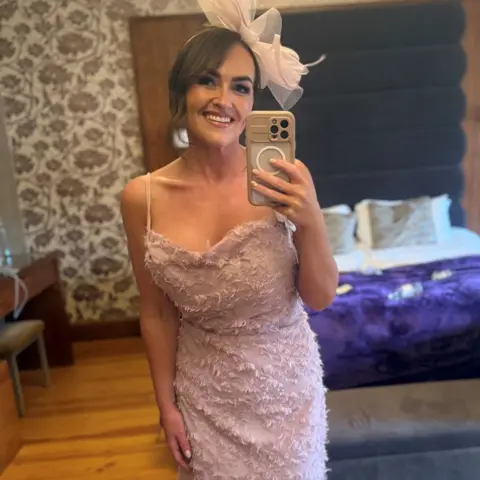 Emilly Murray @itsanewmeemj A woman in a pink dress and fascinator stands in front of a bed, smiling, as she takes a mirror selfie