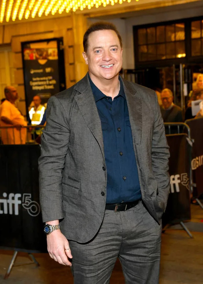Person in a textured suit with a dark shirt, smiling at an event with bright lighting in the background