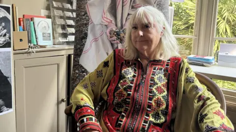 BBC/Jo Burn Deidre Kashdan sitting inside her studio. She is wearing a yellow and red oversized shirt.