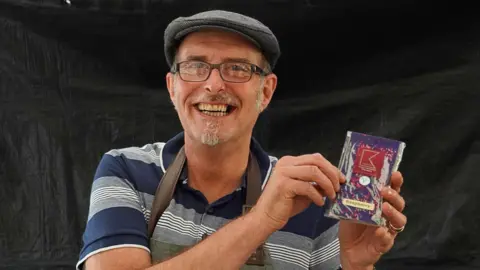 Moseley Farmers' Market A man in a flat cap, who is wearing a navy blue striped polo top is holding a small box of chocolates and smiling at the camera. 