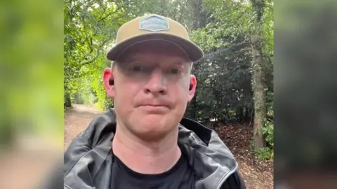 Men Only IOW Aaron Oliver looking at the camera is wearing a beige cap and black waterproof jacket, has a woodland in the background.