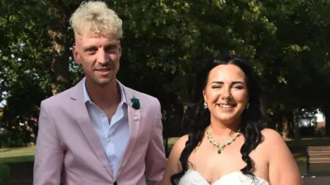 Sammi Hill Sammi Hill and her husband Adam Hill. Sammi has black hair which is partially tied back. She is wearing a strapless wedding dress with a gold and green necklace. Adam is standing on the left, wearing a baby pink suit. He is taller than Sammi and has blonde curly hair with short sides. They are standing in front of a large tree. 