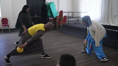 Three women pictured in traditional Muslim dress. One is trying to throw a ball past another to score a goal. 