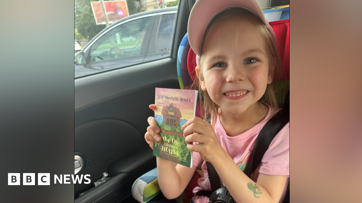 A young girl sits in a car seat smiling at the camera as she holds a book. She is wearing a pink cap and a pink T-shirt. A green fake tattoo is on her forearm in the shape of an octopus.