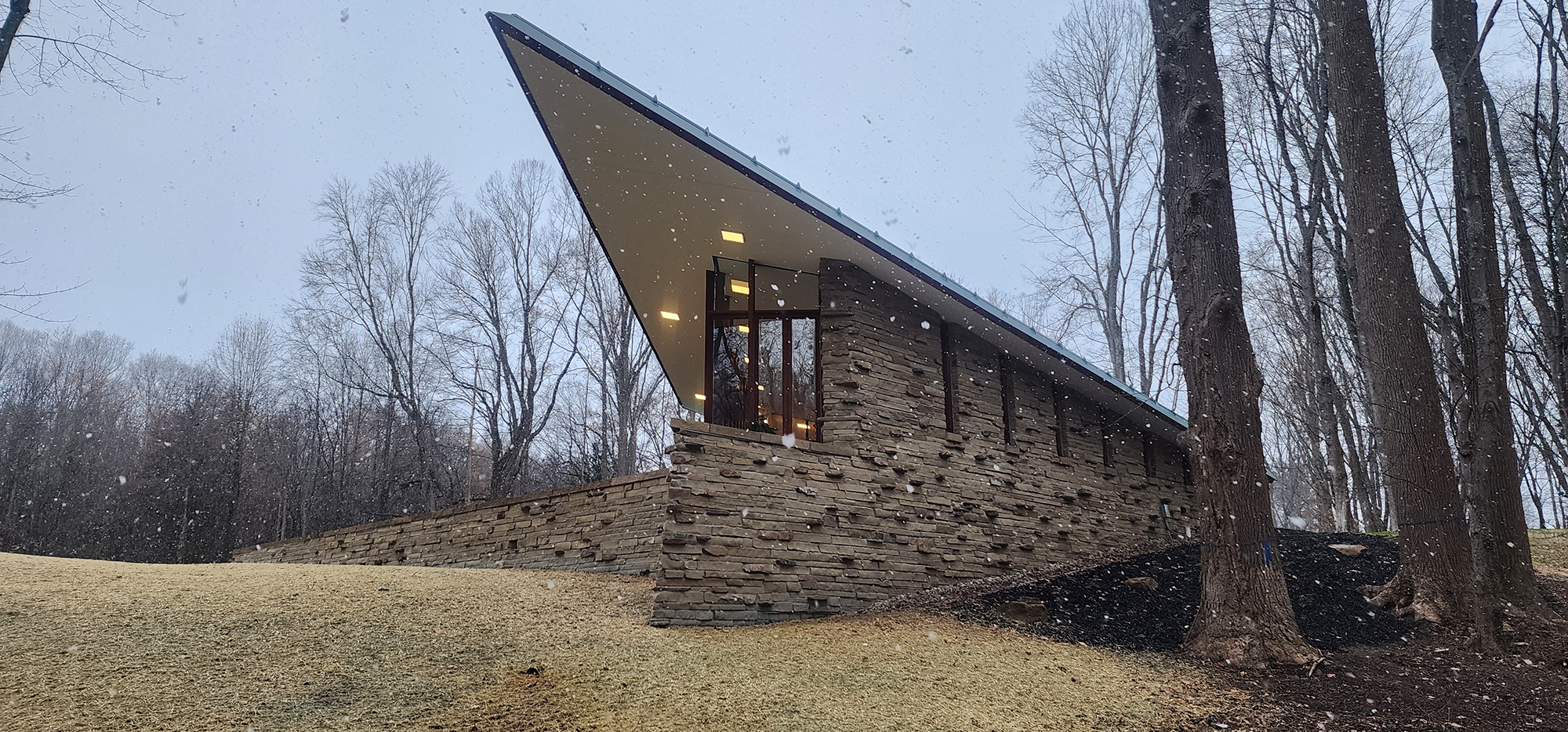 Modern house with a sharply angled roof, stone exterior, and large windows, situated in a snowy, wooded landscape with bare trees.