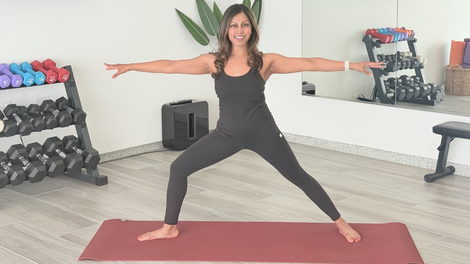 woman in black vest and leggings performing a warrior two pose facing the camera on a red mat in a studio setting. there's a mirror behind her and a rack of weights on the side.