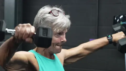 Shaun Whitmore/BBC Fran Smith is pictured lifting a dumbbell in each arm away from her body. She has short grey hair, glasses that sit on top of her head, and she wears a bright green vest top. 