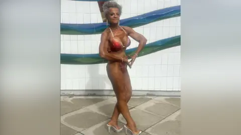 Contributed Ms Smith is pictured prior to a bodybuilding competition. She wears a red bikini and silver heels and she has fake tanned her body. She poses and flexes her arms to show off the muscle definition. 