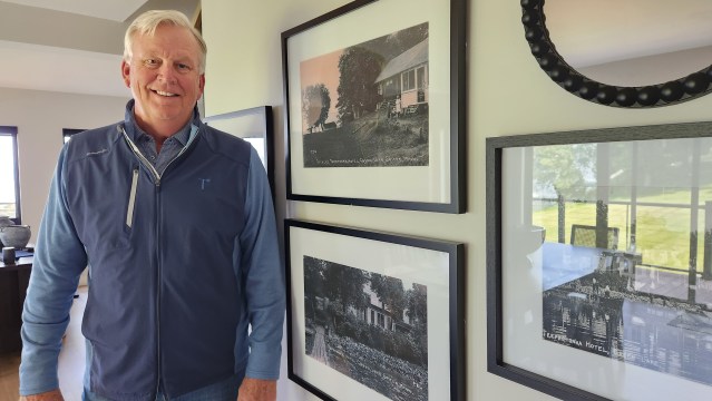 A man with white hair in a blue vest stands next to a wall hung with large framed photos.