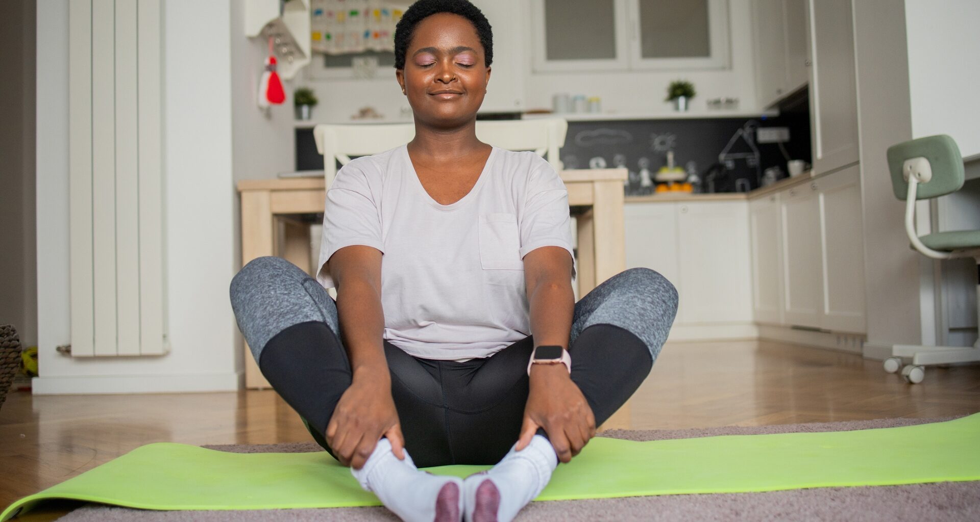 woman wearing a white tshirt and leggings sitting on the floor on a green yoga mat in butterfly pose, facing the camera with eyes closed. there's a kitchen behind her.