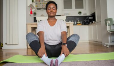 woman wearing a white tshirt and leggings sitting on the floor on a green yoga mat in butterfly pose, facing the camera with eyes closed. there's a kitchen behind her.