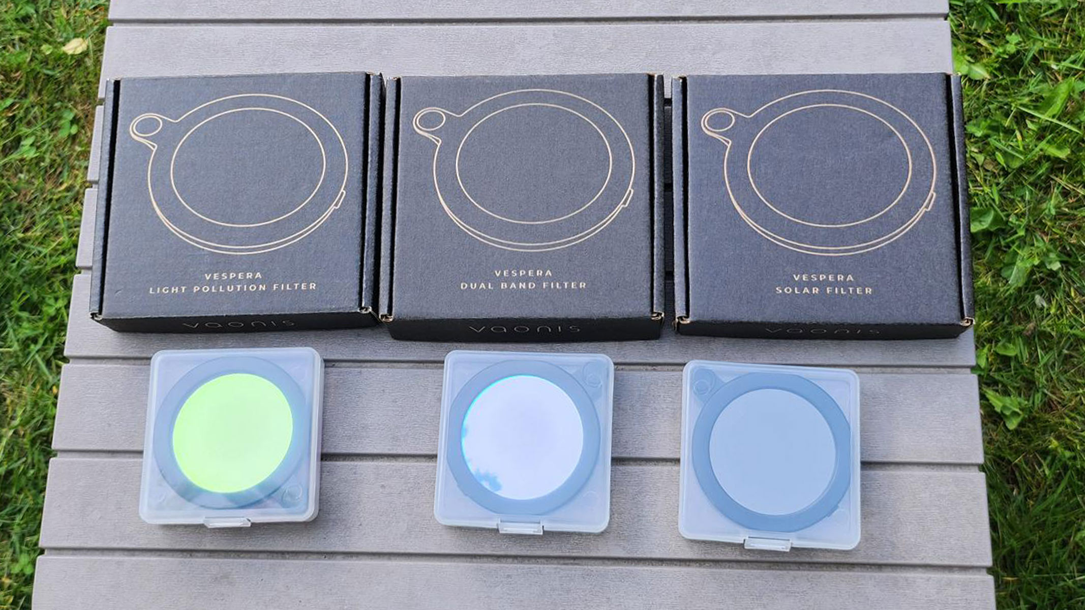 The Vespera Light Pollution Filter, Vespera Dual Band Filter and Vespera Solar Filter with boxes on a grey table with grass at the side.