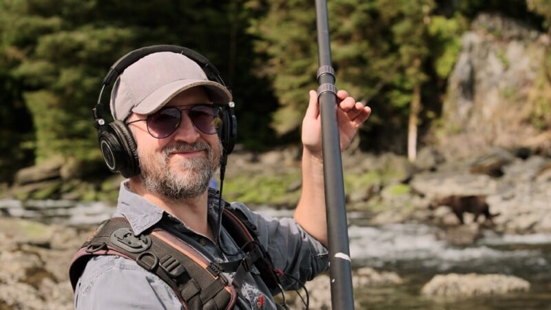 A smiling man wearing headphones, sunglasses, and a cap holds a boom microphone outdoors by a river, with trees, rocks, and a bear in the background.