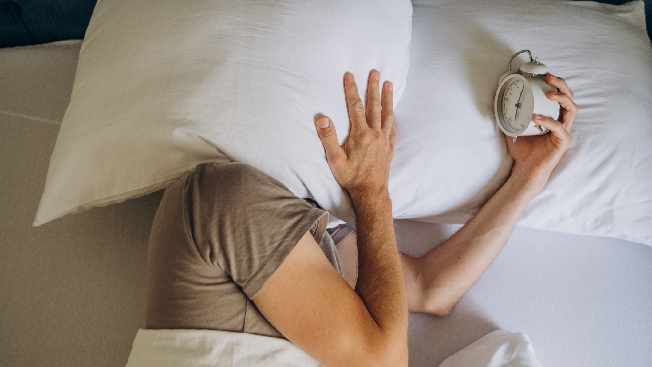 A man who is unable to sleep at night covers his head with a white pillow while holding a white alarm clock in the other hand