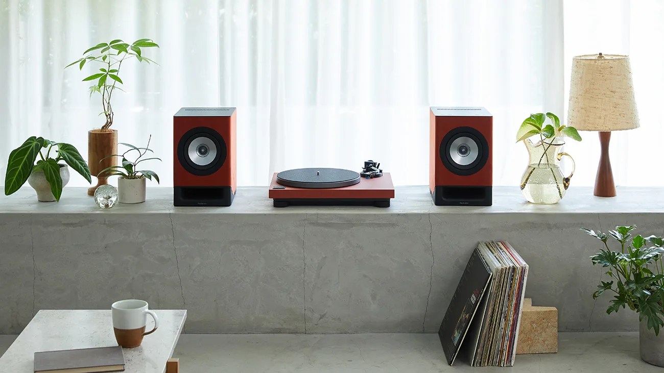 A minimalist setup featuring a red turntable flanked by two matching red speakers on a concrete ledge. The ledge also holds several small potted plants, a glass pitcher with water and green leaves, and a wooden table lamp with a beige shade. Below the ledge, there is a stack of vinyl records leaning against the wall. In the foreground, a white table with a brown and white coffee mug and a closed book is visible. The background consists of sheer white curtains allowing soft natural light.