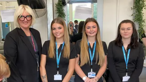 BBC Three young girls standing next to their female college tutor. The tutor has short blonde hair and is wearing glasses with thick black frames. The two girls to her left have long blonde hair. The girl on the end of the line has long dark hair. All of them are wearing black and have lanyards around their necks.
