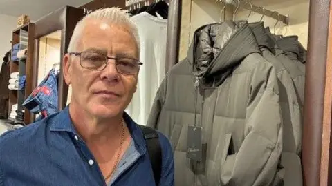 Robert Ryan in a menswear shop with coats and tops on hangers and shelves behind him.