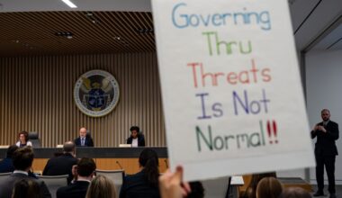 A person attending a Federal Communications Commission meeting holds a sign that says, "governing thru threats is not normal!!"