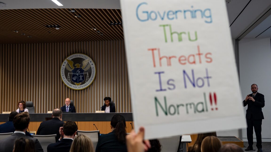 A person attending a Federal Communications Commission meeting holds a sign that says, "governing thru threats is not normal!!"