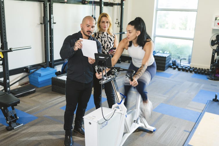 man and woman in street clothes show a paper to a woman in exercise gear on stationary bike