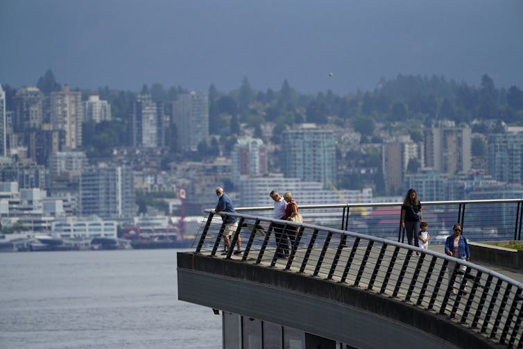 people walk along a waterfront, buildings in the background on the opposing shore