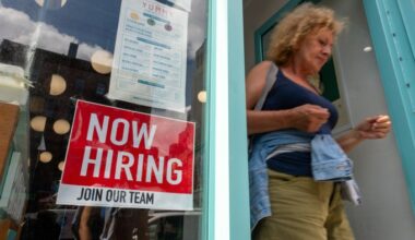 A "Now Hiring, Join Our Team" sign is displayed in a business window as a woman walks out the door