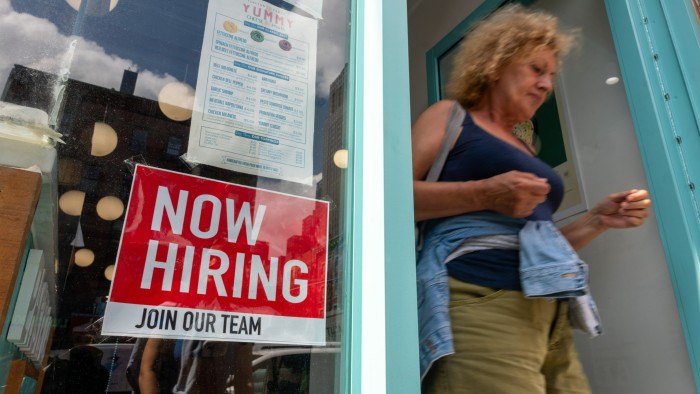 A "Now Hiring, Join Our Team" sign is displayed in a business window as a woman walks out the door
