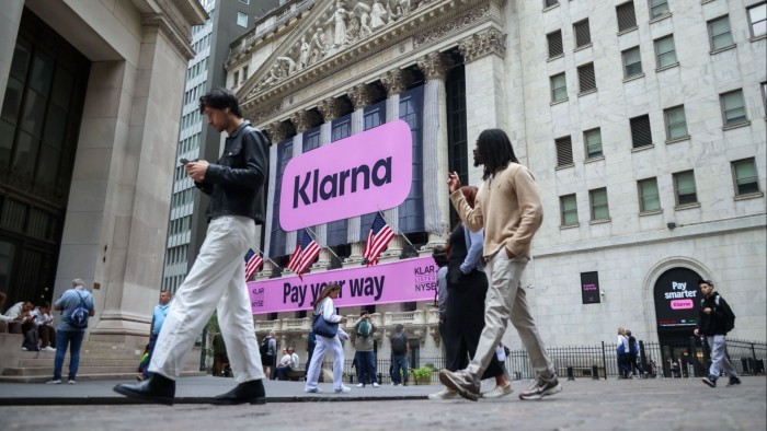 Large pink Klarna signs displayed on the New York Stock Exchange building as people walk by during Klarna's IPO.