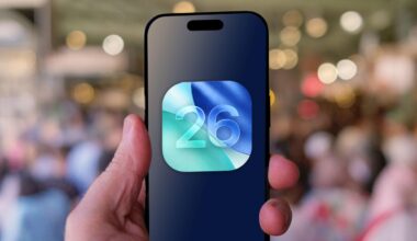 A hand holds an iPhone above a crowd. The logo for iOS 26 is on the screen.